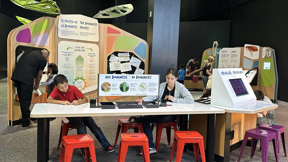 A boy and woman sit on the other side of a table in a science exhibit with people and other exhibit pieces in the back